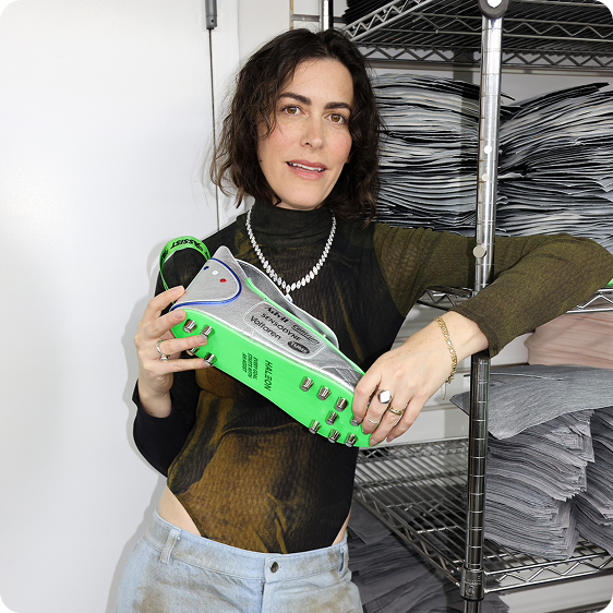 Designer Andrea Bergart poses against a shelf while holding a custom blue, silver, and green dopp kit shaped like a soccer cleat.