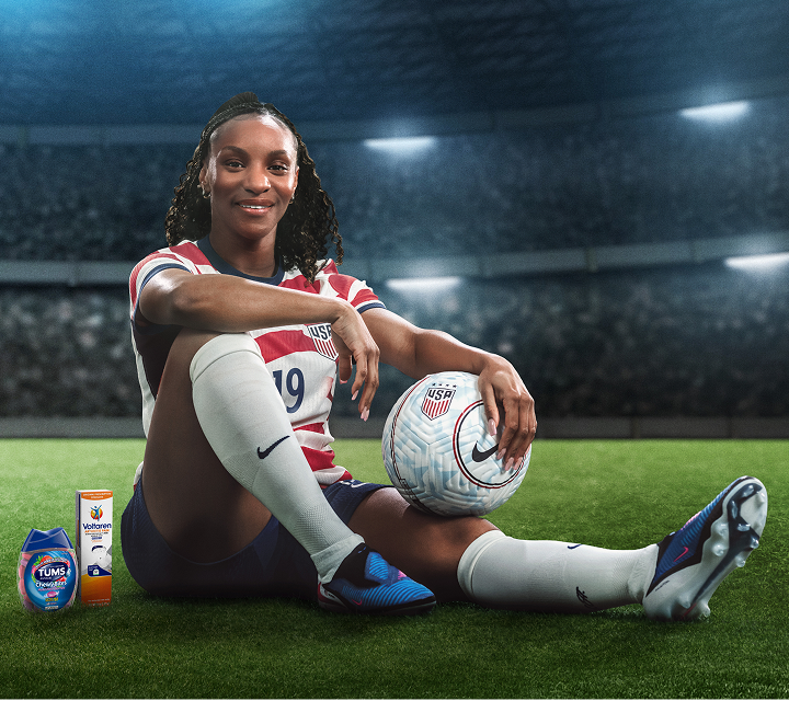 Former U.S. Soccer player Crystal Dunn sits on a field with her back against a goalpost. A ball sits on her knee, while TUMS and Voltaren products are next to her.