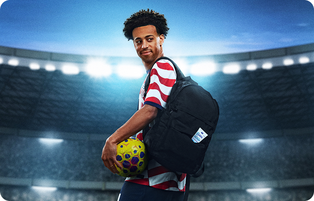 U.S. Soccer player Tyler Adams stands in the middle of a stadium. He holds a soccer ball and a bag with a Pronamel package in the pocket.