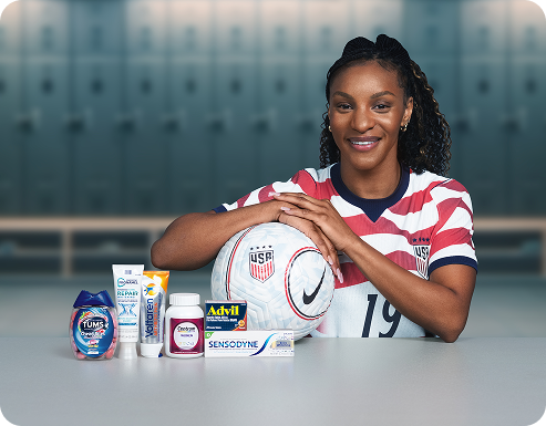 Former U.S. Soccer player Crystal Dunn sits in a locker room with a ball and several Haleon products. Disclaimer says Some products may contain NSAIDs and/or Acetaminophen and should not be taken together. Please review labels and use as directed.