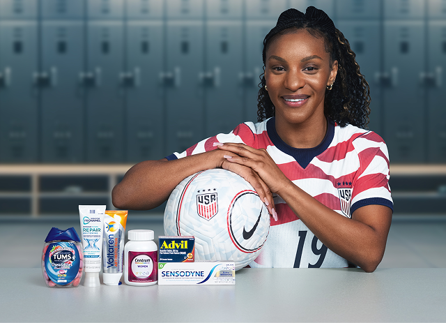 Former U.S. Soccer player Crystal Dunn sits in a locker room with a ball and several Haleon products. Disclaimer says Some products may contain NSAIDs and/or Acetaminophen and should not be taken together. Please review labels and use as directed.