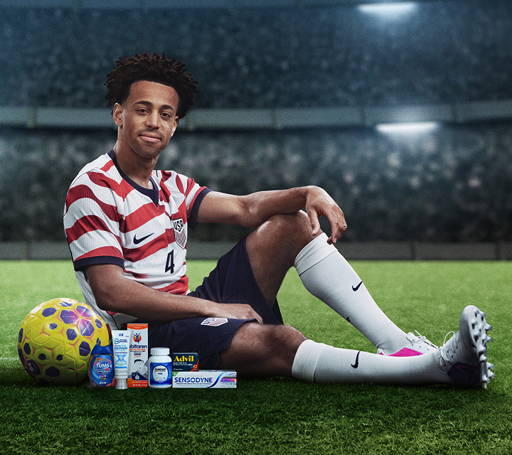 U.S. Soccer player Tyler Adams sits on a soccer field next to a ball and several Haleon products. Disclaimer says Some products may contain NSAIDs and/or Acetaminophen and should not be taken together. Please review labels and use as directed.