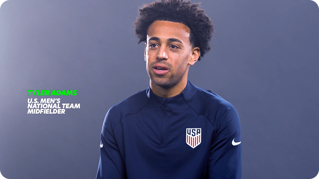 U.S. Soccer player Tyler Adams sits in his warm-up gear, speaking directly to the camera. An overlay with a play video button appears in the corner.